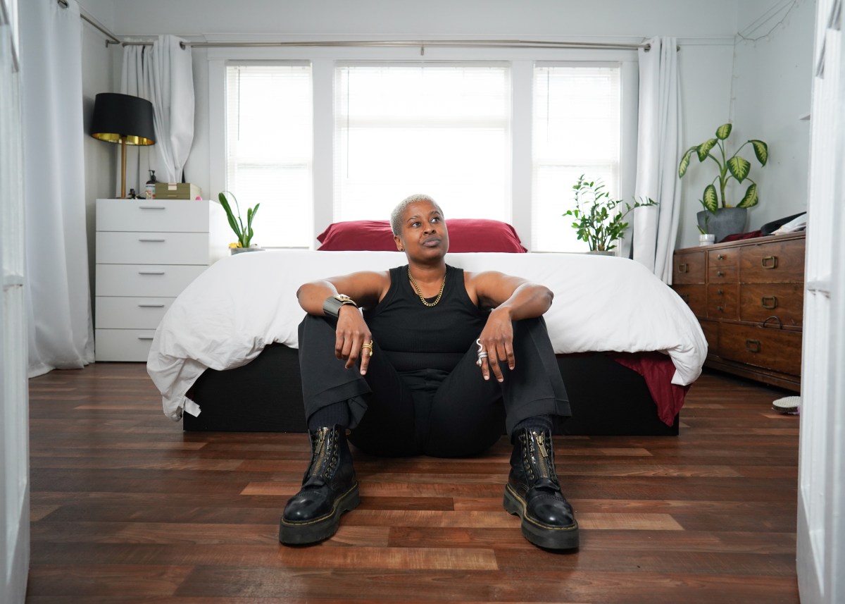 a Black stud sits at the end of a bed, wearing ablack tank and black pants and black boots