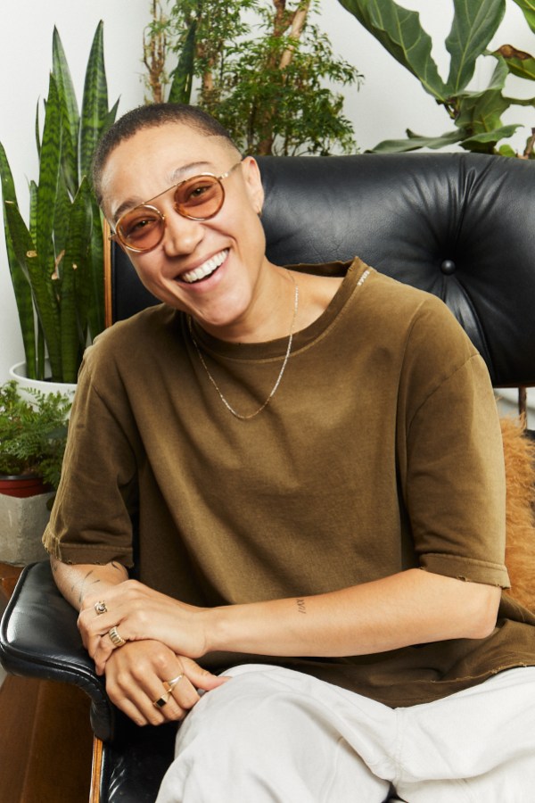 a photo of Kylo Freeman. they are a Black masc of center person with short cut hair and a fade. they are smiling and wearing rose tinted sunglasses, a gold chain, a brown tee, loose white pants and various rings. they are leaning to the side of a chair