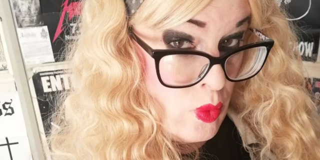 Kat Shevil Gillham, a transwoman metal rocker in the UK, poses in a record store for a selfie. She has blonde hair, a headband, and black frame glasses and is wearing black eyeshadow and red lipstick.