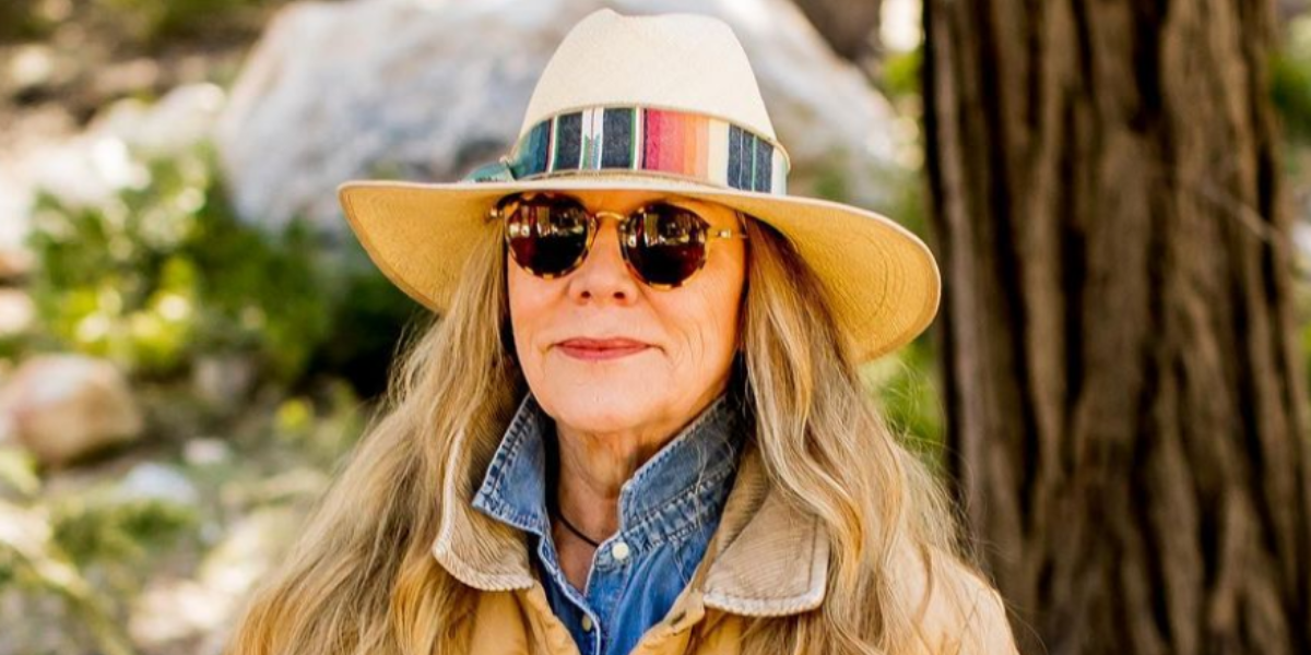 Laura Ann Carleton wearing a wide brimmed hat with a multicolored stripe on it and a jean shirt and camel colored field jacket and sunglasses