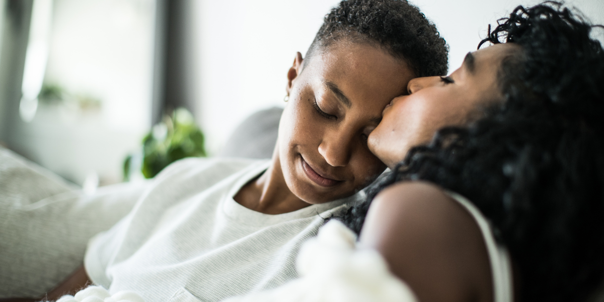 two Black women sit on a couch, and one kisses the other's forehead in a tender moment