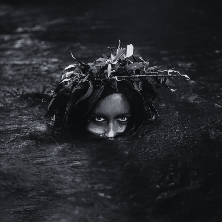a woman looks out from dark waters, her face painted and leaves in her hair