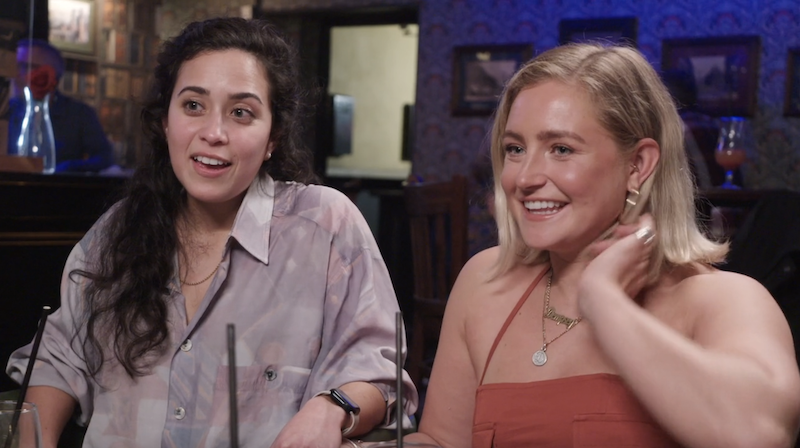 statler on the left wearing a loose button down and smirking. dempsey sitting on her right wearing a burnt orange spaghetti strap top and smiling at her friends off-screen