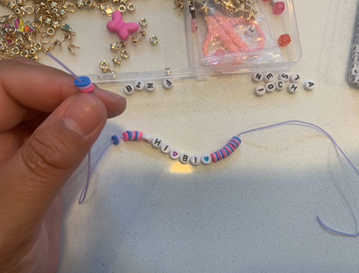 Fingers holding the end of a string with blue, pink, and purple clay beads on it and the words HI BI spelled in letter beads