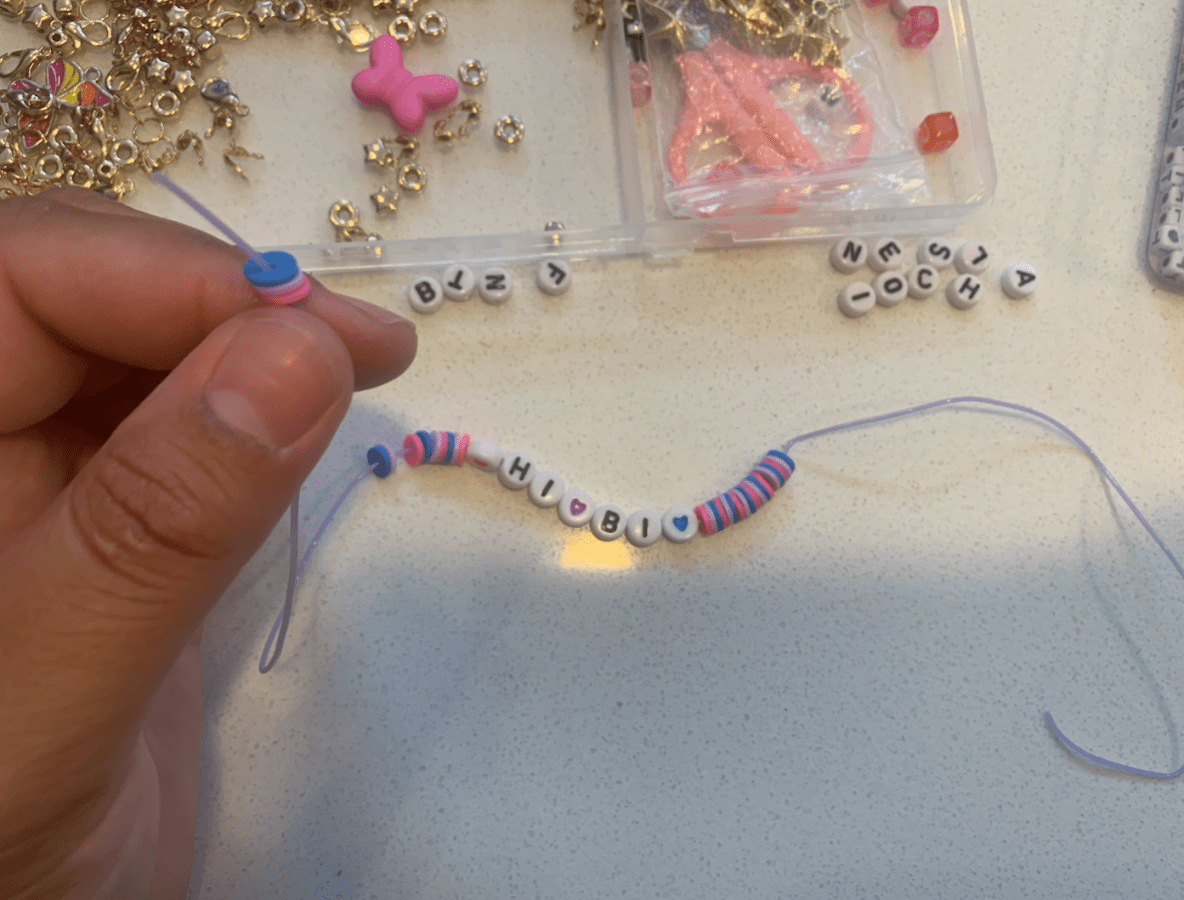 Fingers holding the end of a string with blue, pink, and purple clay beads on it and the words HI BI spelled in letter beads