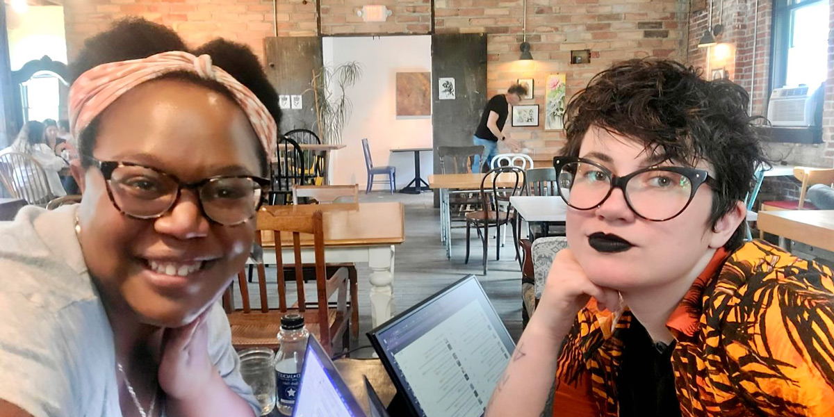 Carmen, a Black woman with glasses, and Nico, a white brunette genderqueer person also with glasses, are smiling for a camera over a table with two laptops open.
