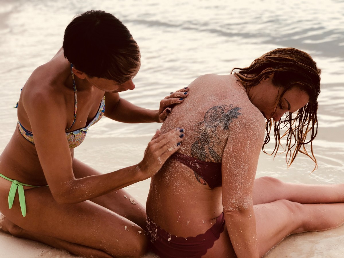 a woman rubs sand on another woman's back on the shore