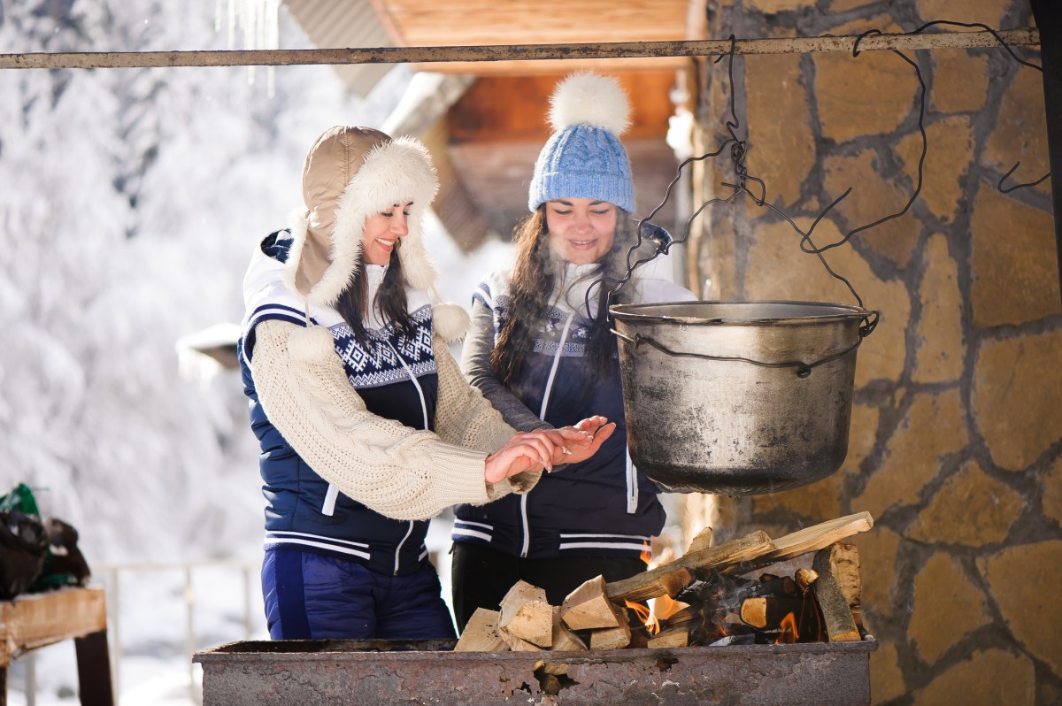 two women bundled in sweaters and furry hats cook a vat of soup over an open flame