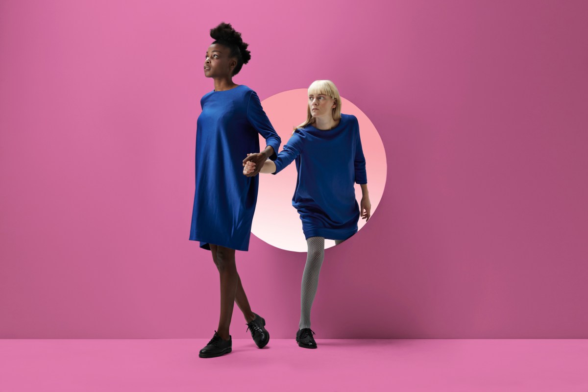 a Black woman leads a white woman by the hand through a circle cut in a pink wall