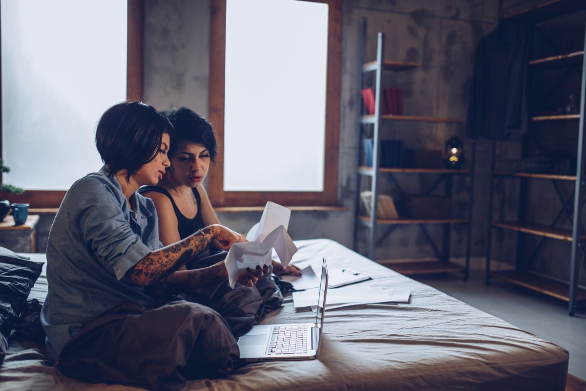 two lesbians sit on their bed doing personal finances on a laptop and looking at receipts and bills