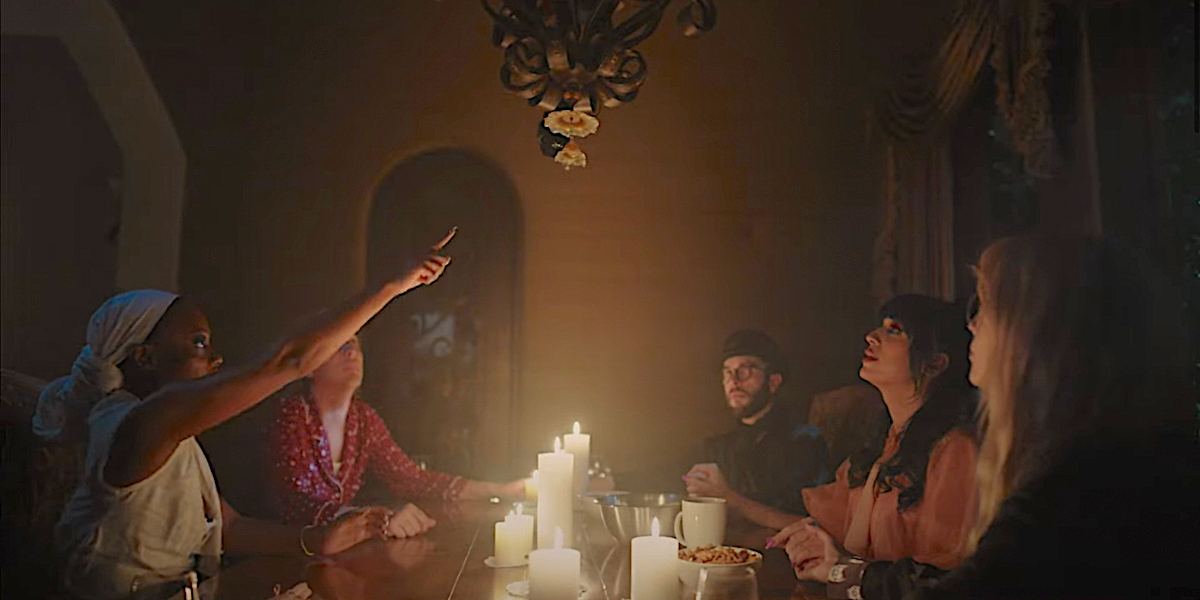 A group of queers sit around a candle lit seance table. One of them points up at a chandelier.