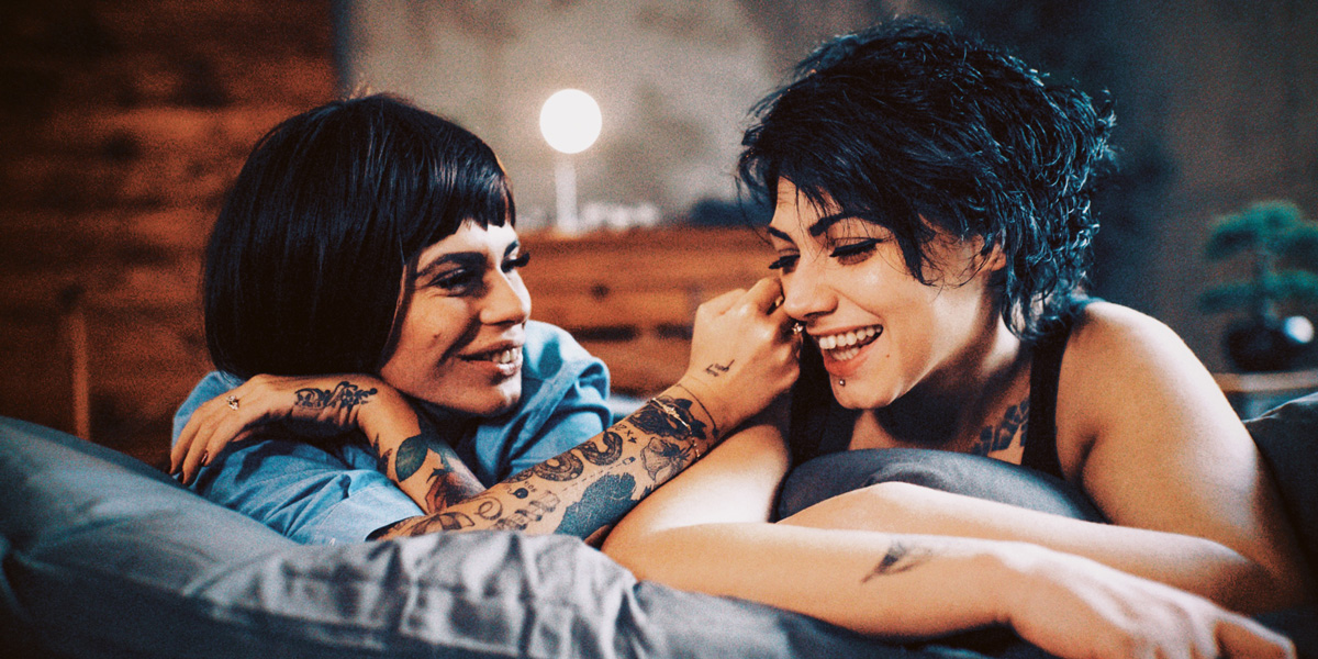 Two white queer humans with tattoos and short brunette hair, one in a pixie cut and one with bangs, caress and smile with each other in bed.