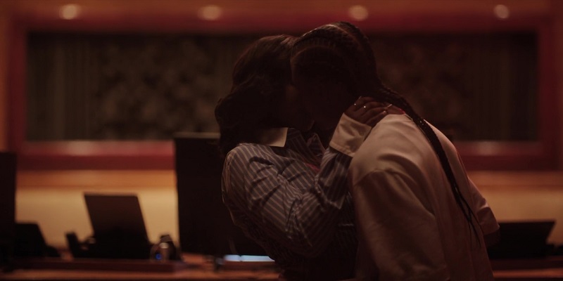 After resisting at first, Jemma pulls Brittany back into a second kiss. The girls are in the studio, standing in front of a row of monitors. Jemma is on the left, wearing a blue and white striped button down with white cuffs. Brittany has on an oversized white tee.
