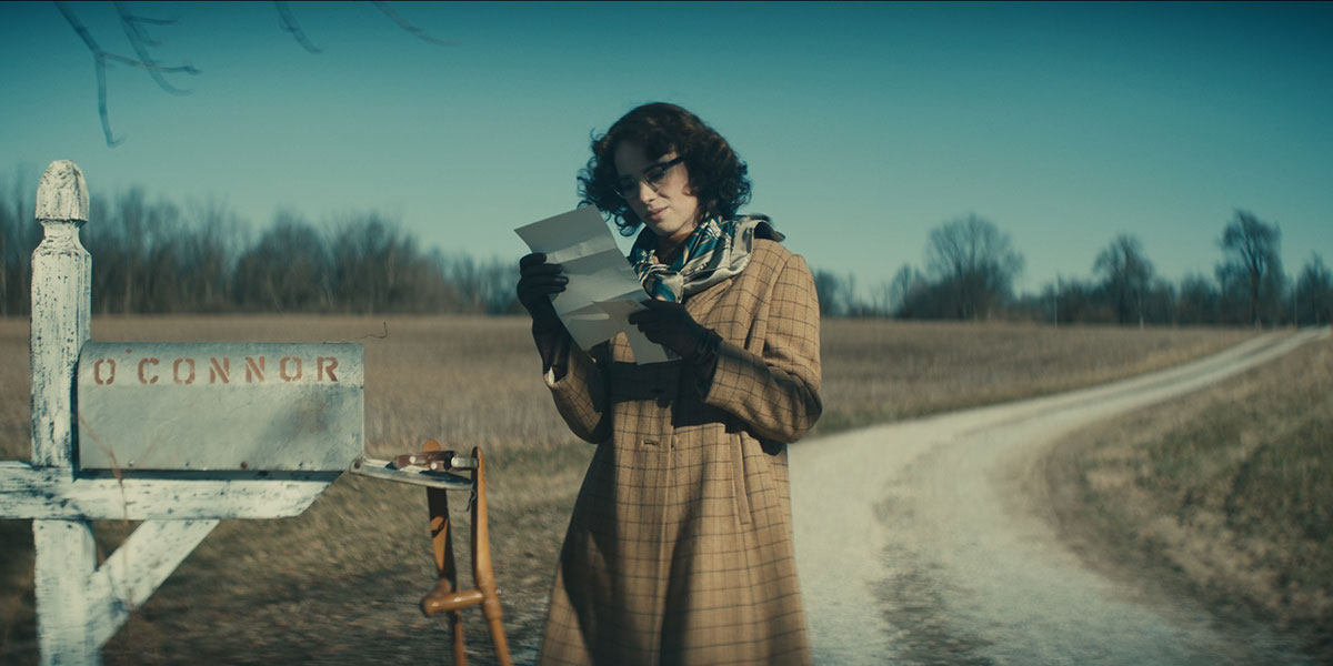 Maya Hawke as Flannery O'Connor reads a letter by a mailbox wearing a brown coat.