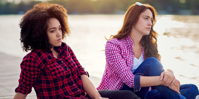 a Black woman wearing a red and black flannel sits next to a white woman in a pink and white checkered buttondown on a dock on the water, both looking contemplative.