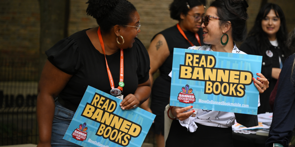 two women look at each other while smiling and holding signs that say READ BANNED BOOKS