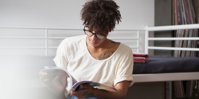 a Black woman with glasses and an off white shirt reads a magazine