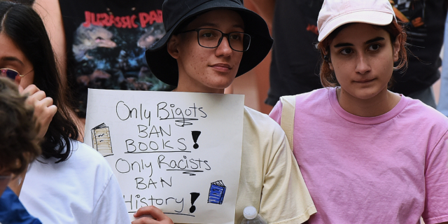 a person holding a sign that says ONLY BIGOTS BAN BOOKS! ONLY RACISTS BAN HISTORY!