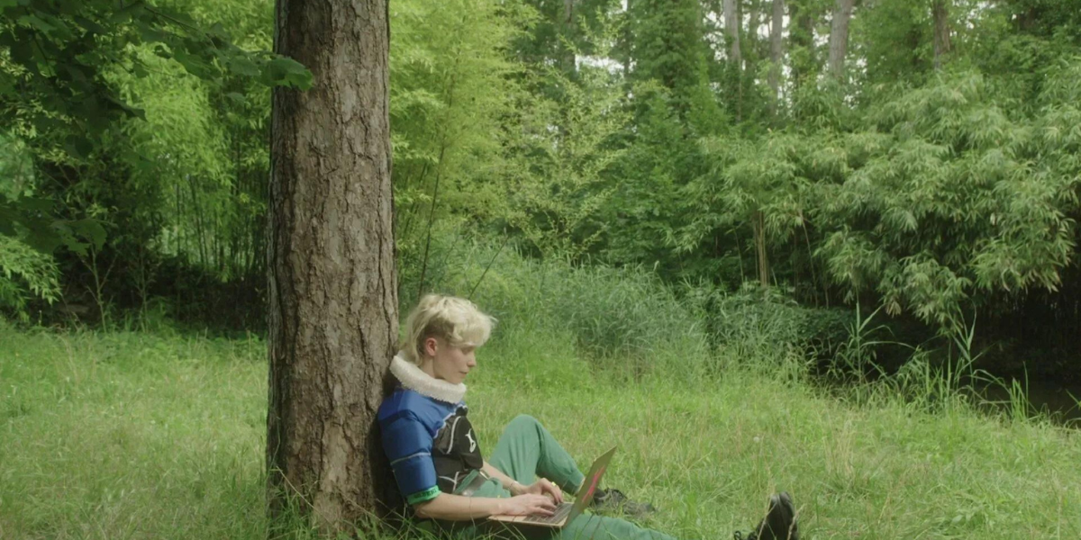 a person with a blonde mullet leans against a tree and types on a laptop in Orlando My Political Biography
