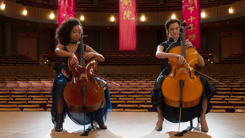 Queer horror to stream: The Perfection. Logan Browning and Allison Williams play cello on stage next to each other with an empty audience behind them.