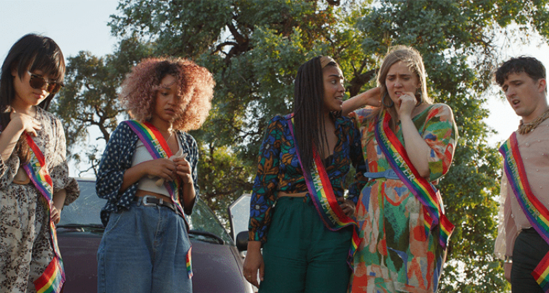 the cast of Sissy stands in rainbow sashes, looking at something on a road