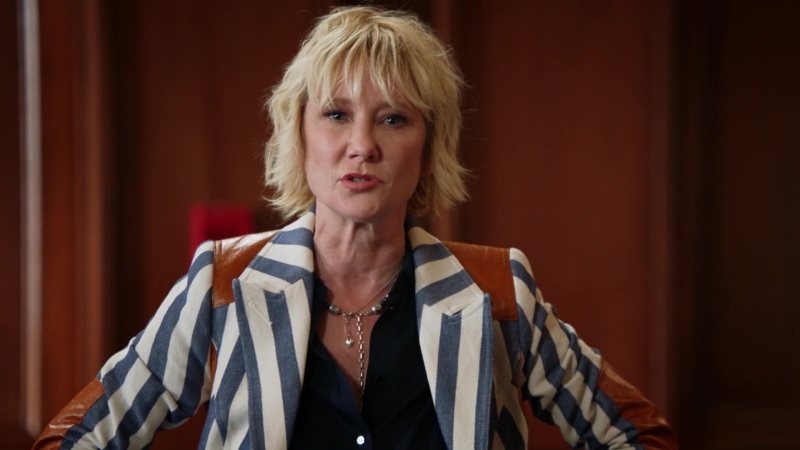 The late Anne Heche as Corrine Cuthbert, addressing the court, wearing an indigo blue striped equestrian blazer with whiskey leather patches.