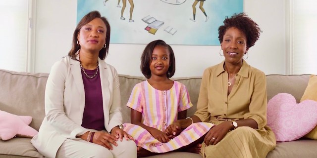 Laphonza Butler (left) sits beside her daughter, Nylah, and her partner, Neneki Lee. They're all sitting on a tan couch in front of a canvas of black girls playing double dutch.