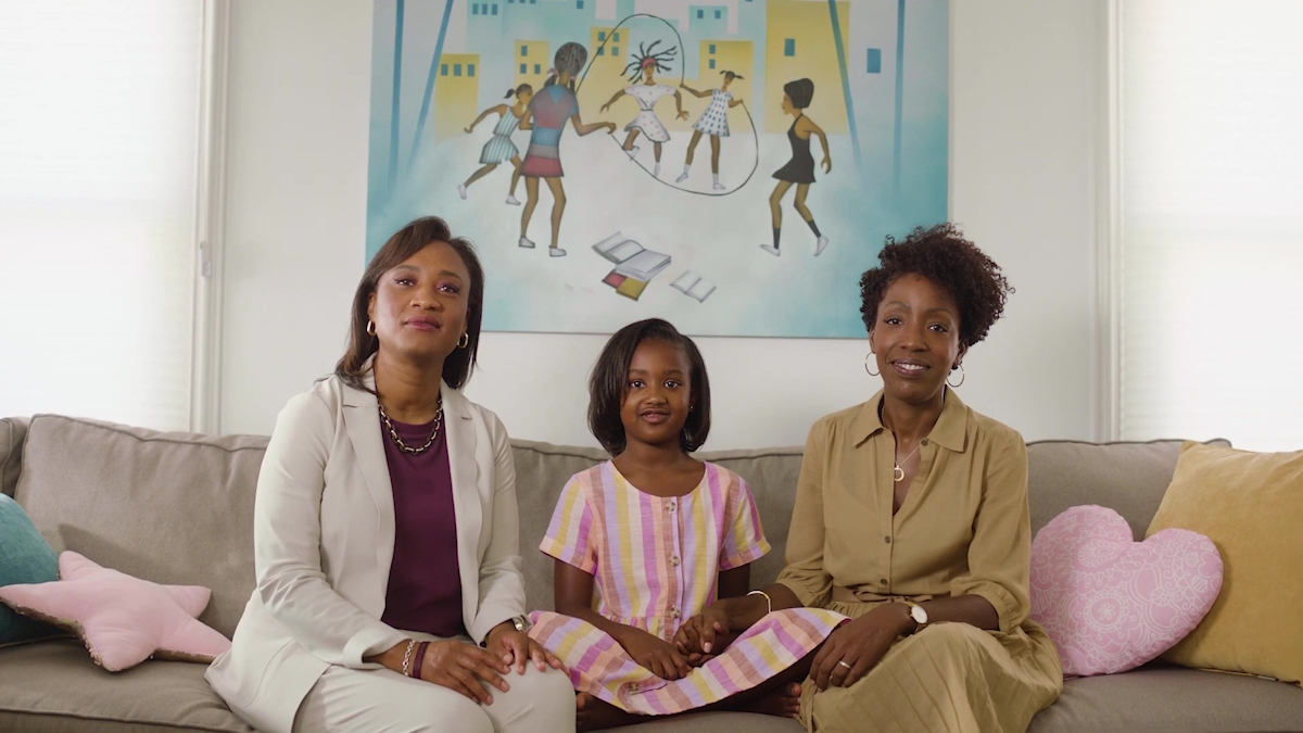 Laphonza Butler (left) sits beside her daughter, Nylah, and her partner, Neneki Lee. They're all sitting on a tan couch in front of a canvas of black girls playing double dutch.