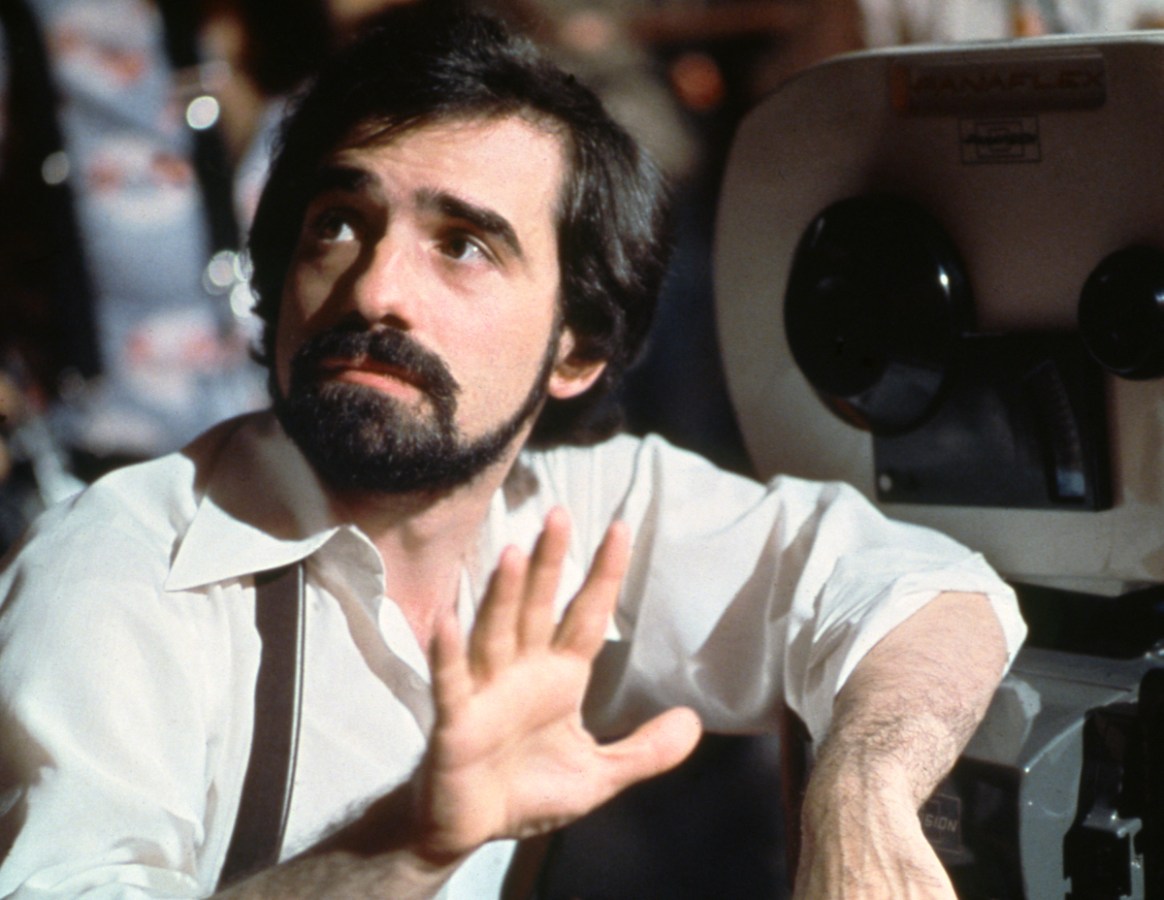 A young Martin Scorsese lifts his hand up and looks off to the side as he leans against a camera.