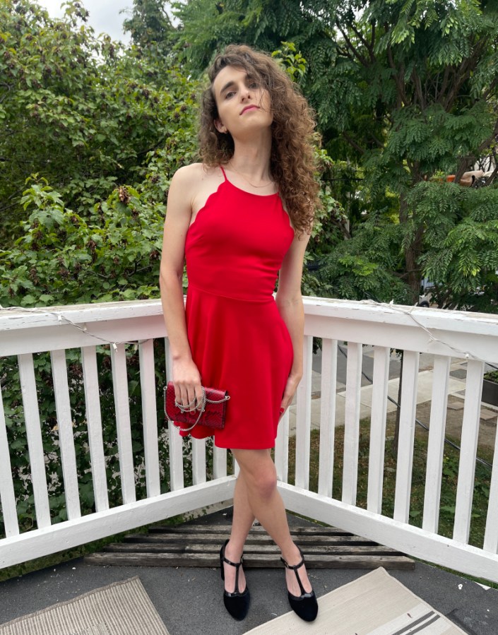 Drew poses on a balcony in a red dress. She clutches a red purse, is wearing black shoes, and has her head tilted up, her hair partially covering half her face. 