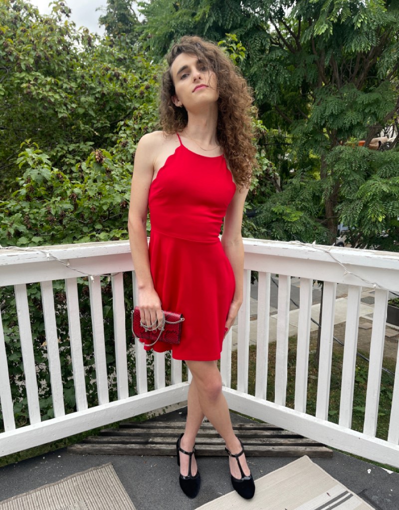 Drew poses on a balcony in a red dress. She clutches a red purse, is wearing black shoes, and has her head tilted up, her hair partially covering half her face. 