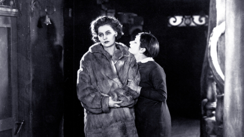 A young Greta Garbo in a fur coat looks forlorn as another woman grabs onto her arm. 