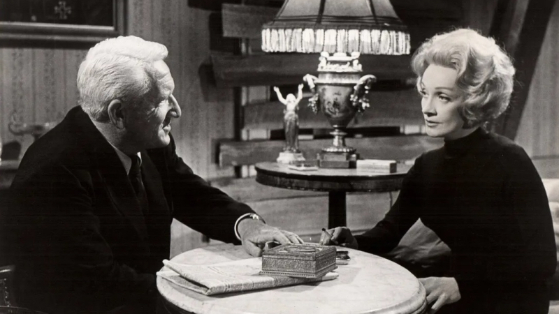 An older Marlene Dietrich dressed in black sits across a table from Spencer Tracy