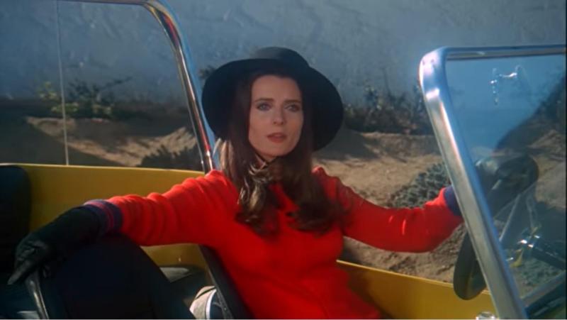 a vampire woman sits in a convertible car, wearing a red dress and black hat in the lesbian vampire movie The Velvet Vampire