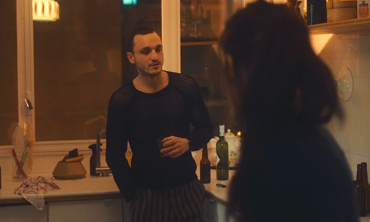 Clothes from Passages: Franz Rogowski in a long sleeve mesh top has a drink in a kitchen while talking to someone.