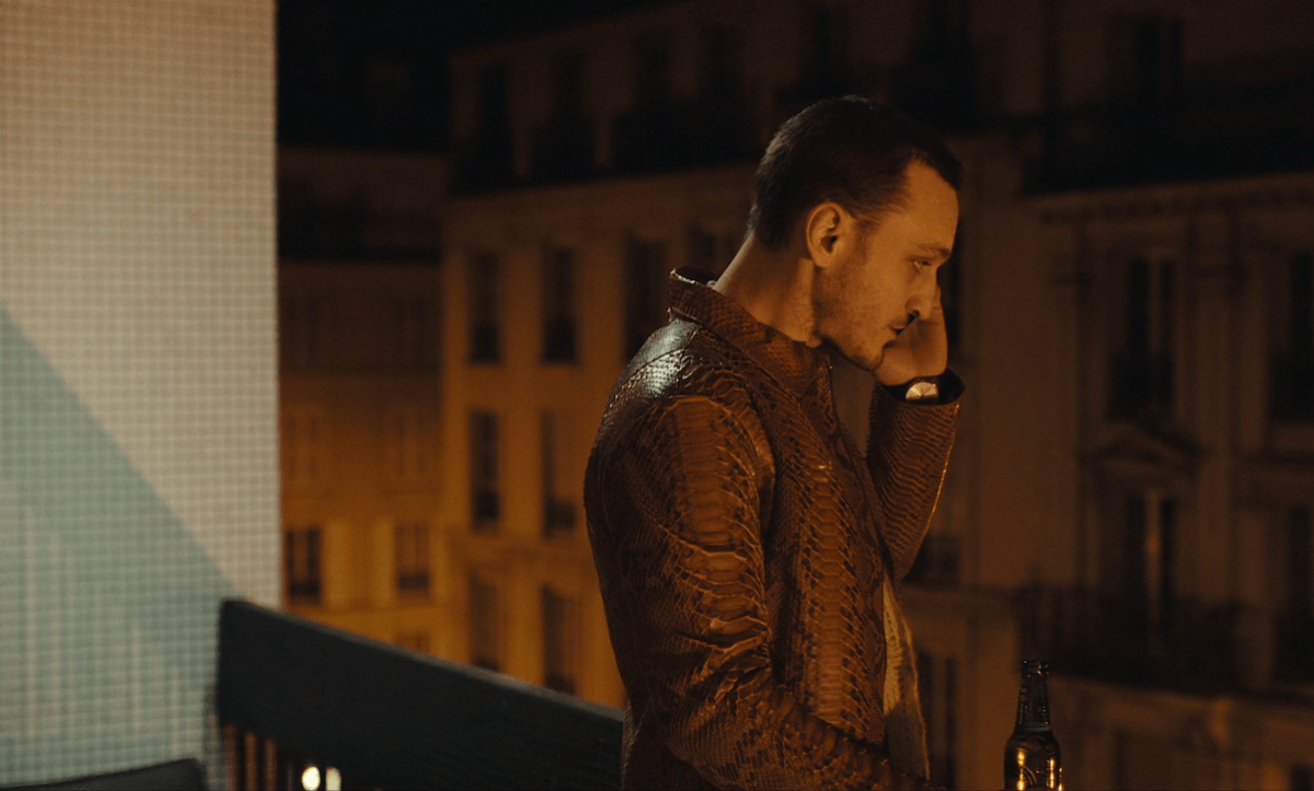 Franz Rogowski wearing a snakeskin jacket stands on a dark balcony talking on the phone.
