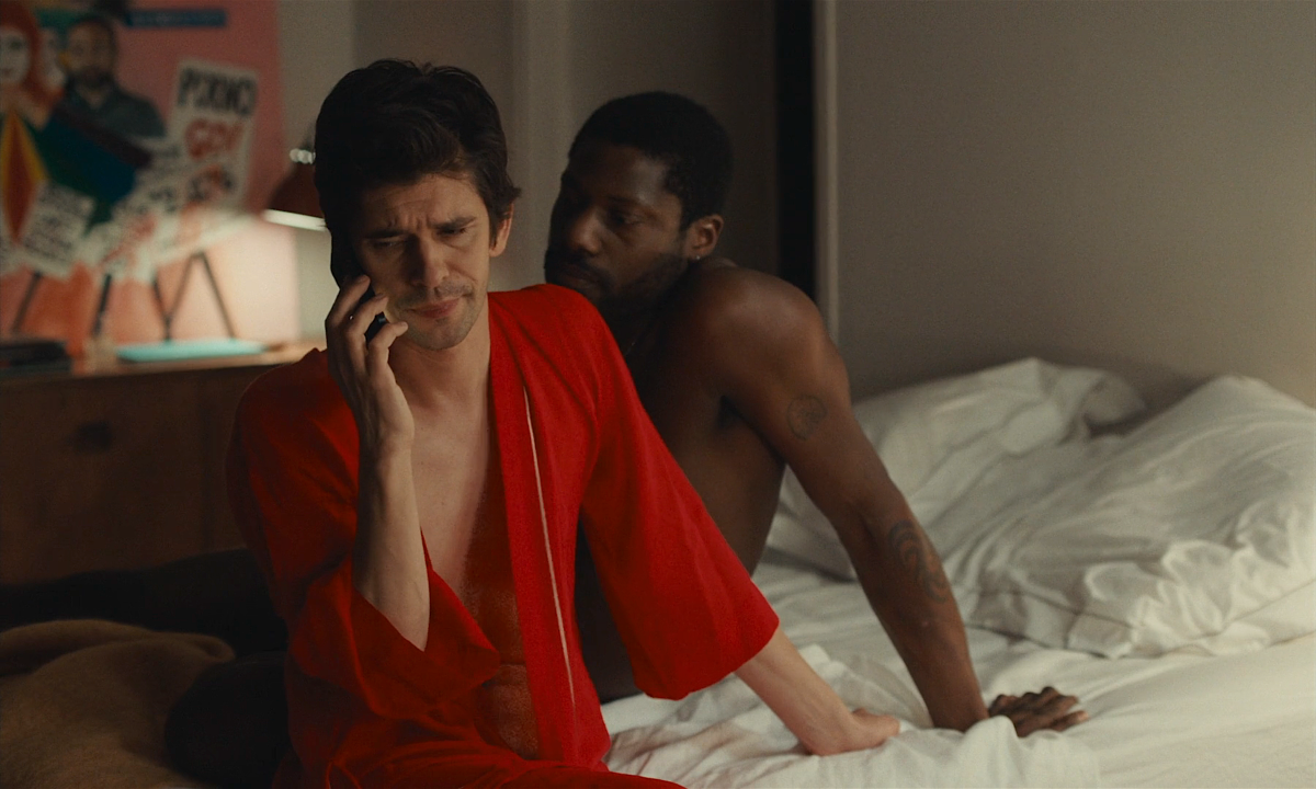 Ben Whishaw in an open red robe talks on the phone. A shirtless man sits up in bed besides him.