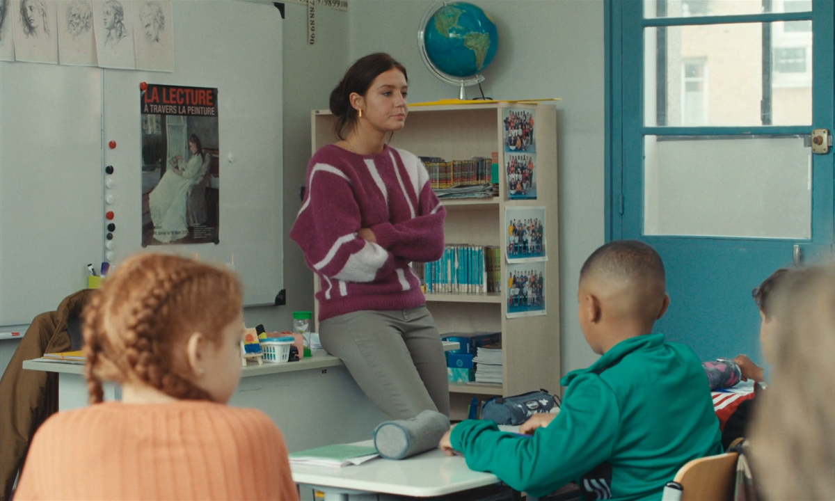 Adèle Exarchopolous in tight light pants and a purple and white sweater leans against a desk in a classroom as children look on.