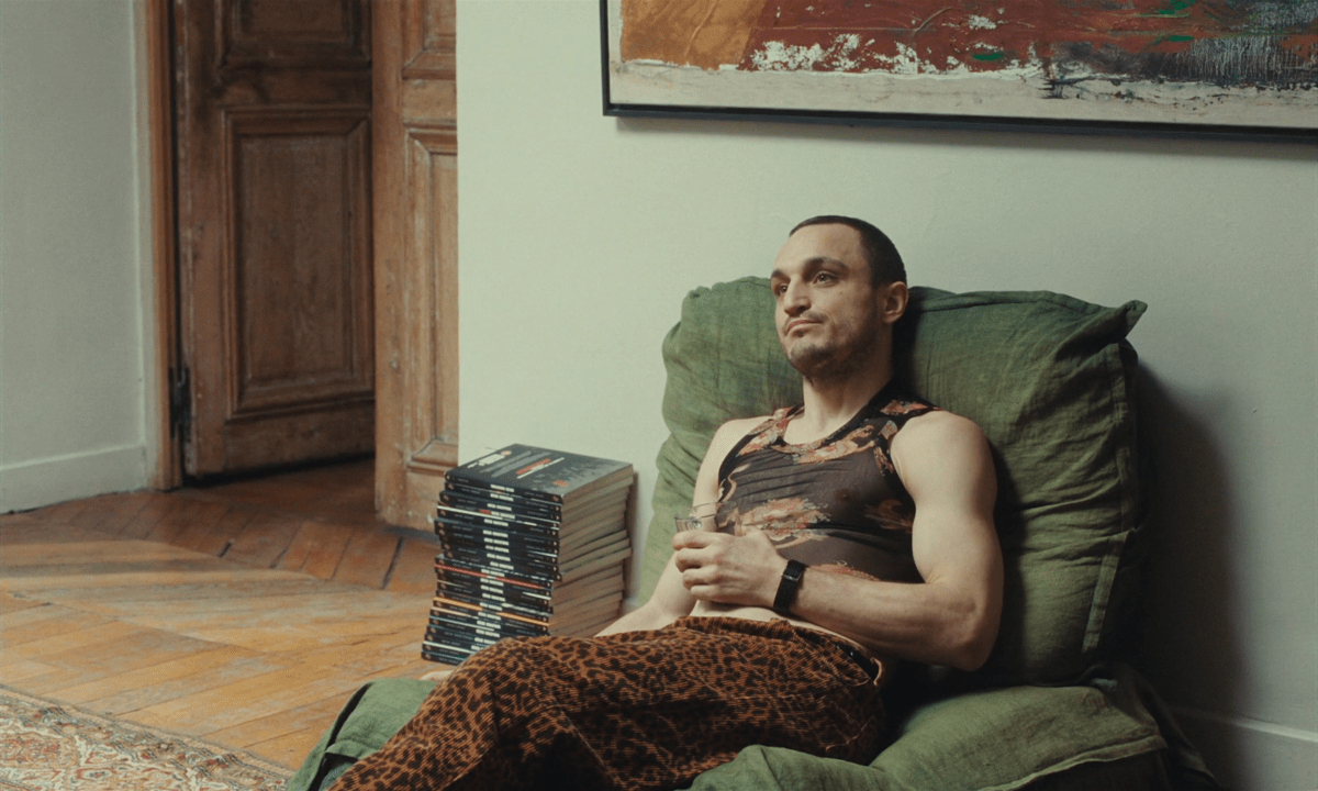 Franz Rogowski sits on a green chair wearing a patterned black sleeveless crop top and loose leopard pants.