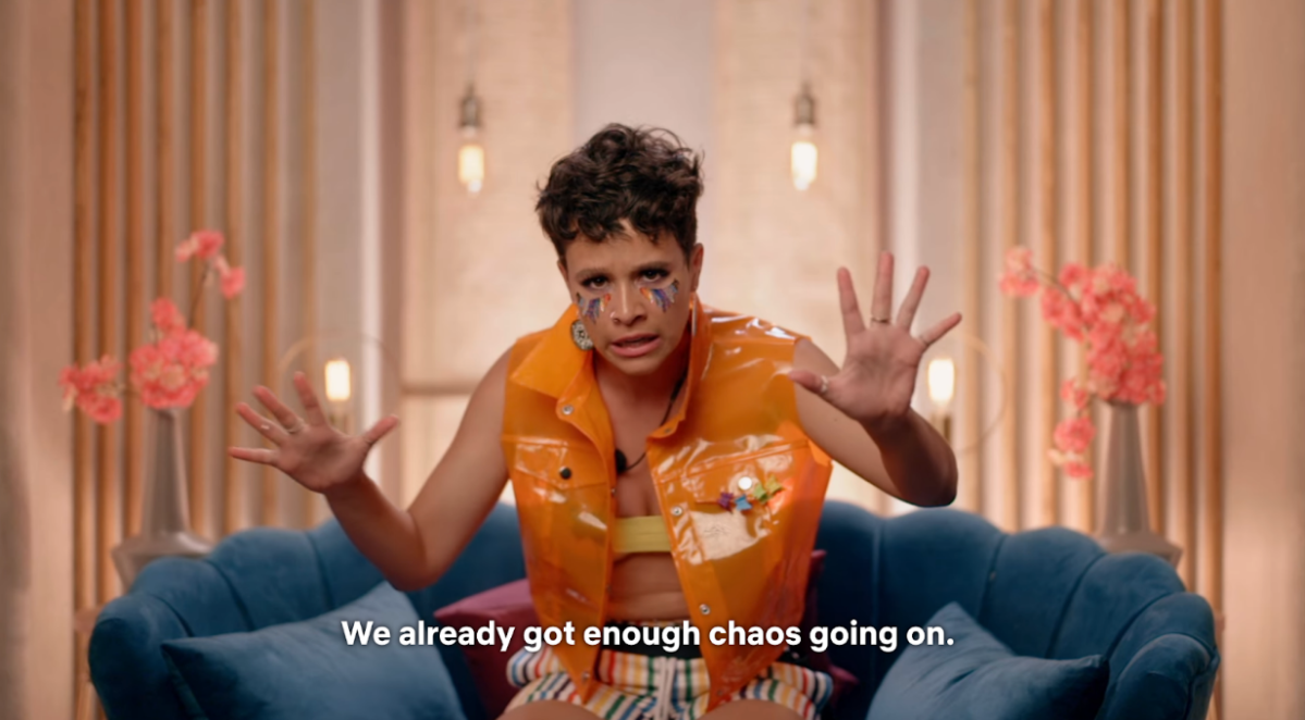 Surviving Paradise contestant Tabitha Sloan in rainbow make-up and a neon orange vest with just a piece of yellow duct tape under it gesticulates wildly, saying “We already got enough chaos going on”