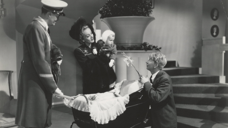 Marlene Dietrich holds a baby as one man has a hand on the carriage and another man holds up a flower.
