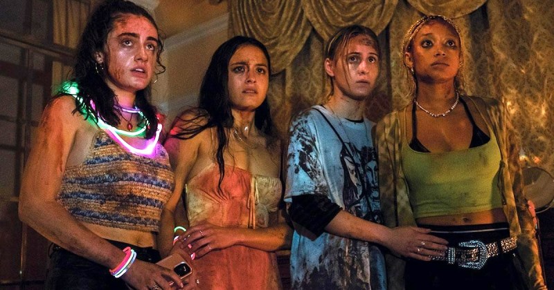 four teens in the movie Bodies Bodies Bodies, covered in blood and sweat and wearing glow in the dark necklaces look at something in horror