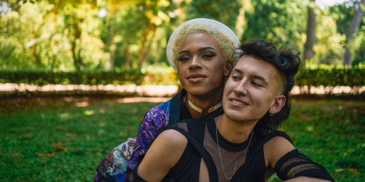 two queer folks in a park, holding onto each other. one is wearing a white hat and patterned track jacket, and one is wearing a black mesh top with cutouts on the shoulders.