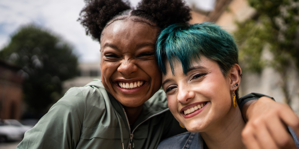 a Black femme and a white person with blue short hair who are both young adults embrace and smile.