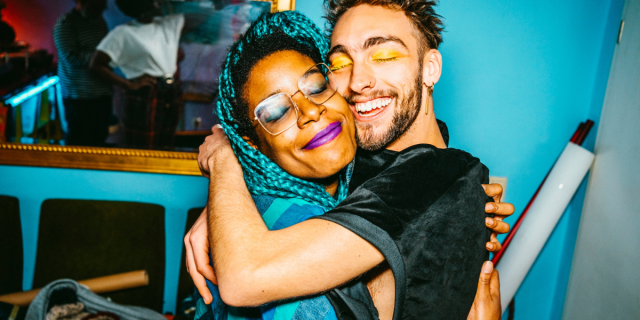 a Black femme with glasses and a purple lip smiles and hugs a white masc person with a beard and gold eye shadow who is also smiling