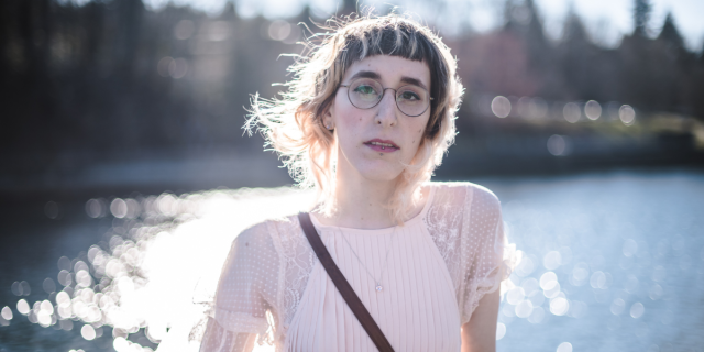 a queer person wearing glasses and a pale pink blouse standing in the sunshine