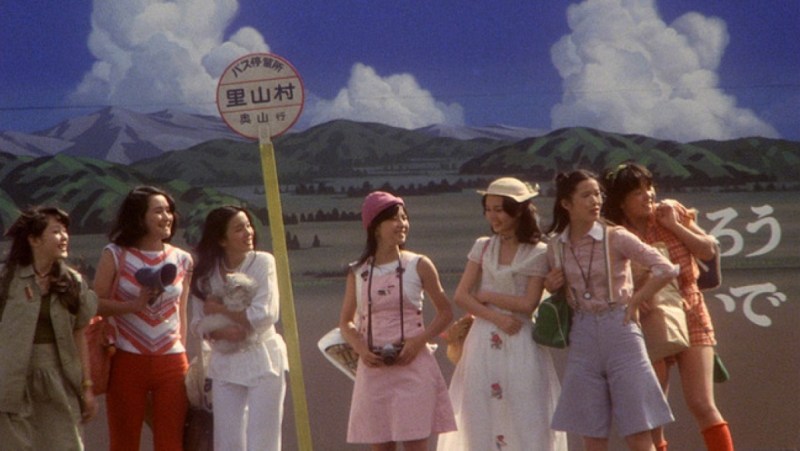 A group of colorfully dressed girls stand close together at a bus stop with a painted skyline behind them.
