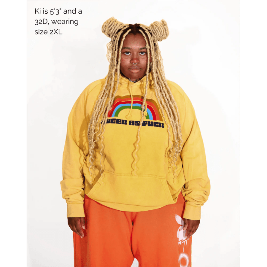 model in a mustard Queer as Fuck hoodie with a rainbow on it
