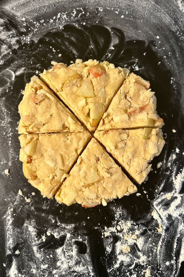 A circle of patted down dough for apple cheddar scones, on a floured black counter top.