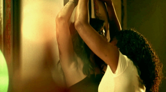 One woman pins another woman against a wall with her arms above her head as they kiss.